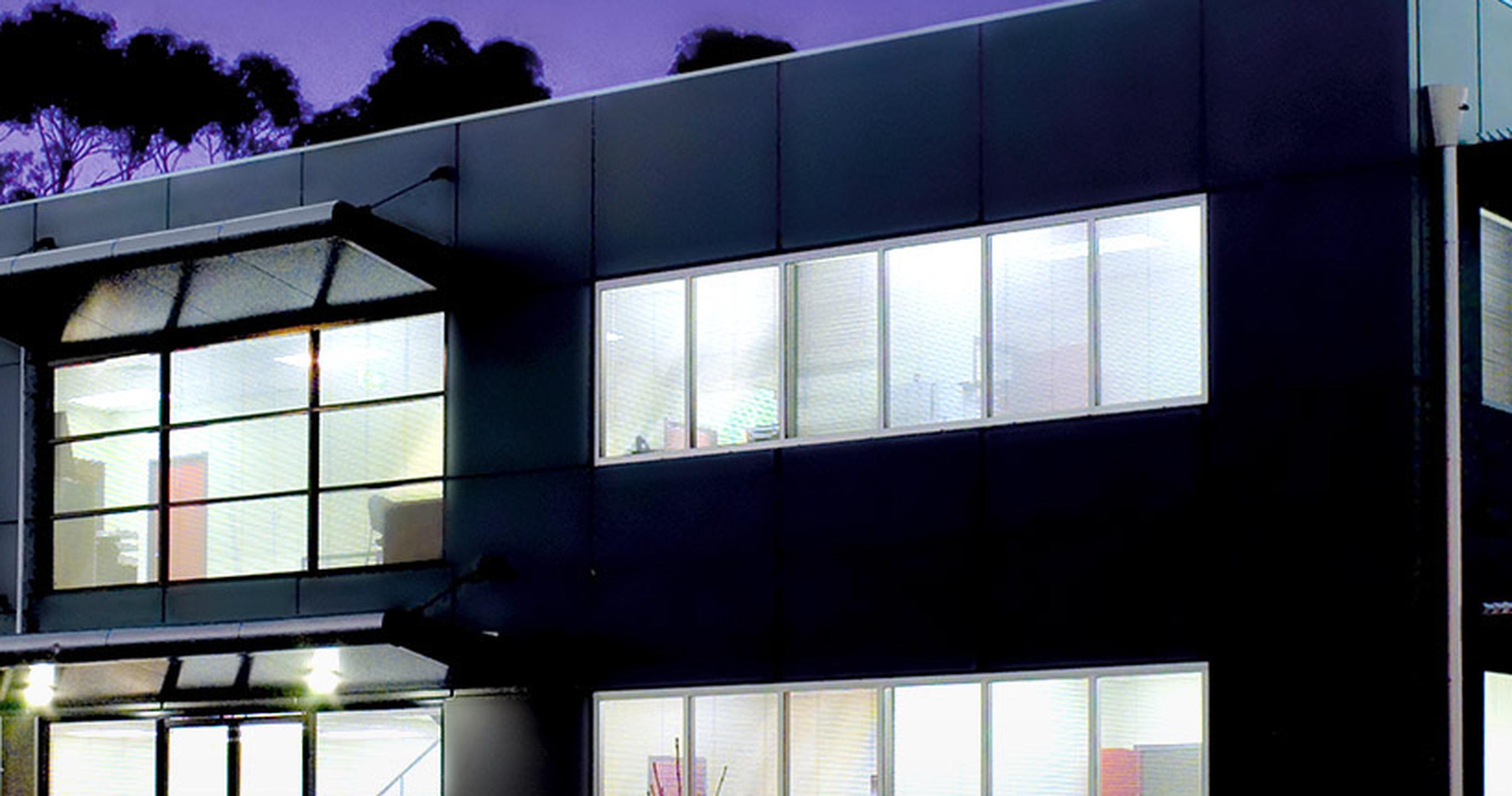 Two-storey dark-clad commercial building at dusk, its office windows lit from within, gum trees behind against a twilight sky